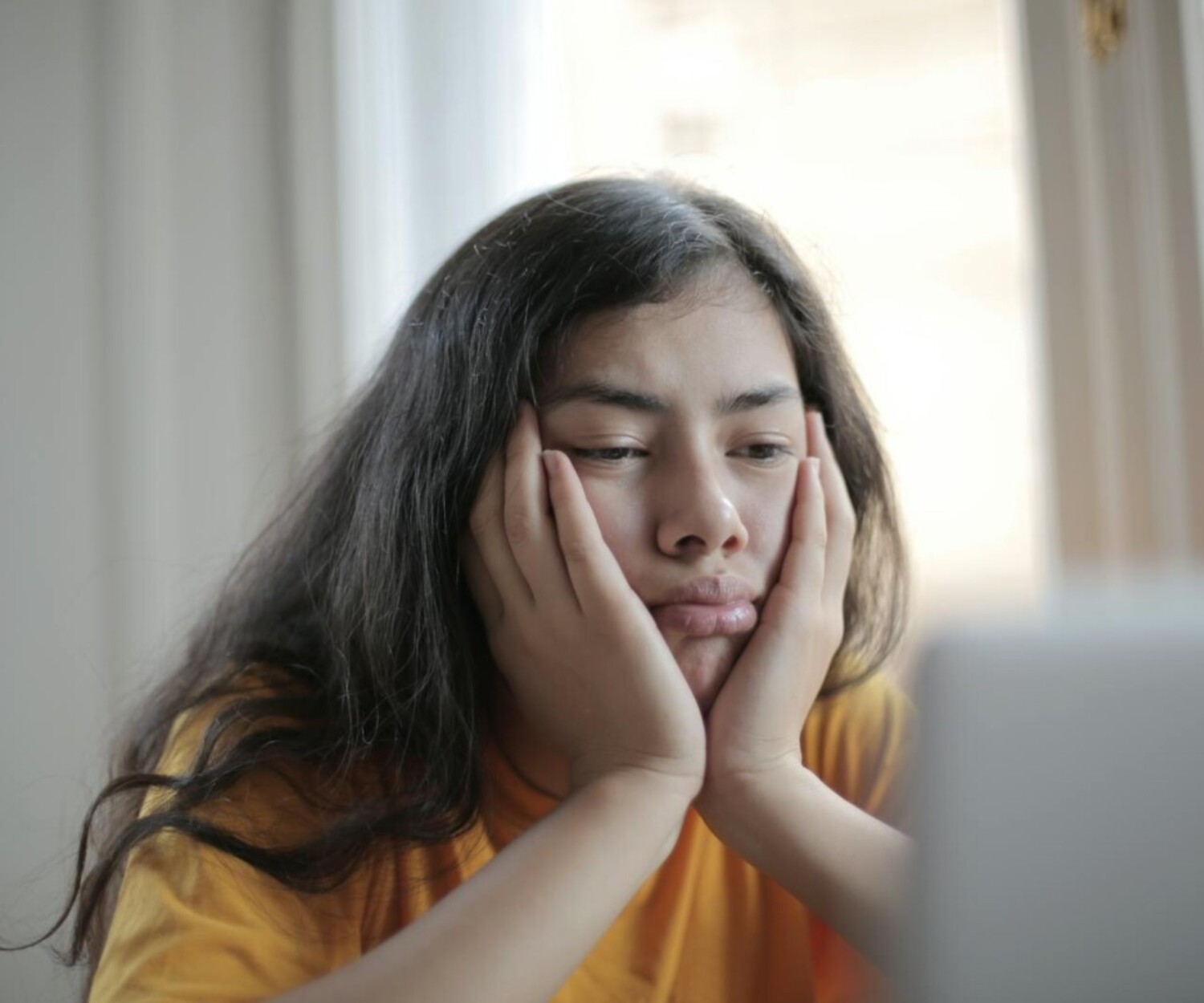 Bored ethnic female in casual clothes sitting at table with netbook and watching sad movie while resting at home during weekend